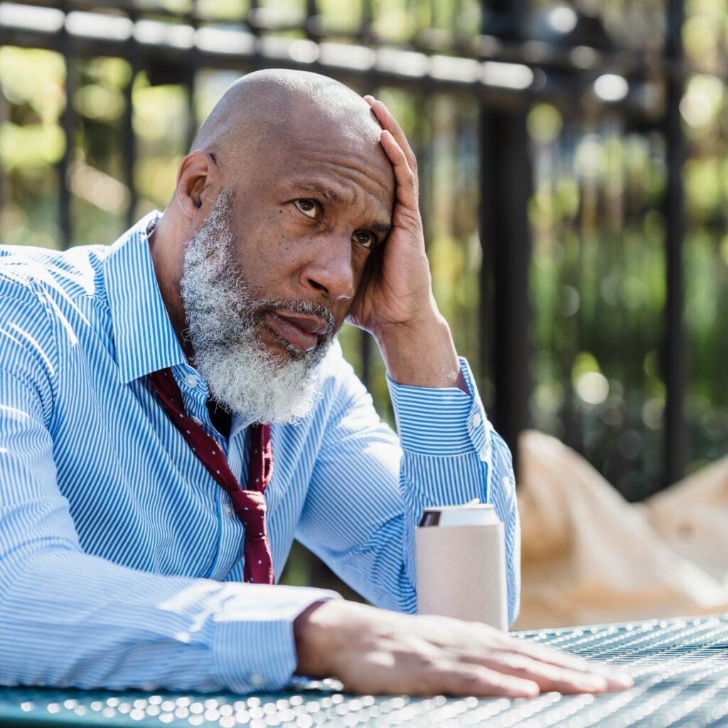 Concentrated senior bearded black male wearing blue shirt and untied tie leaning head on hand while sitting at table with tin can of beverage in veranda cafe in daylight