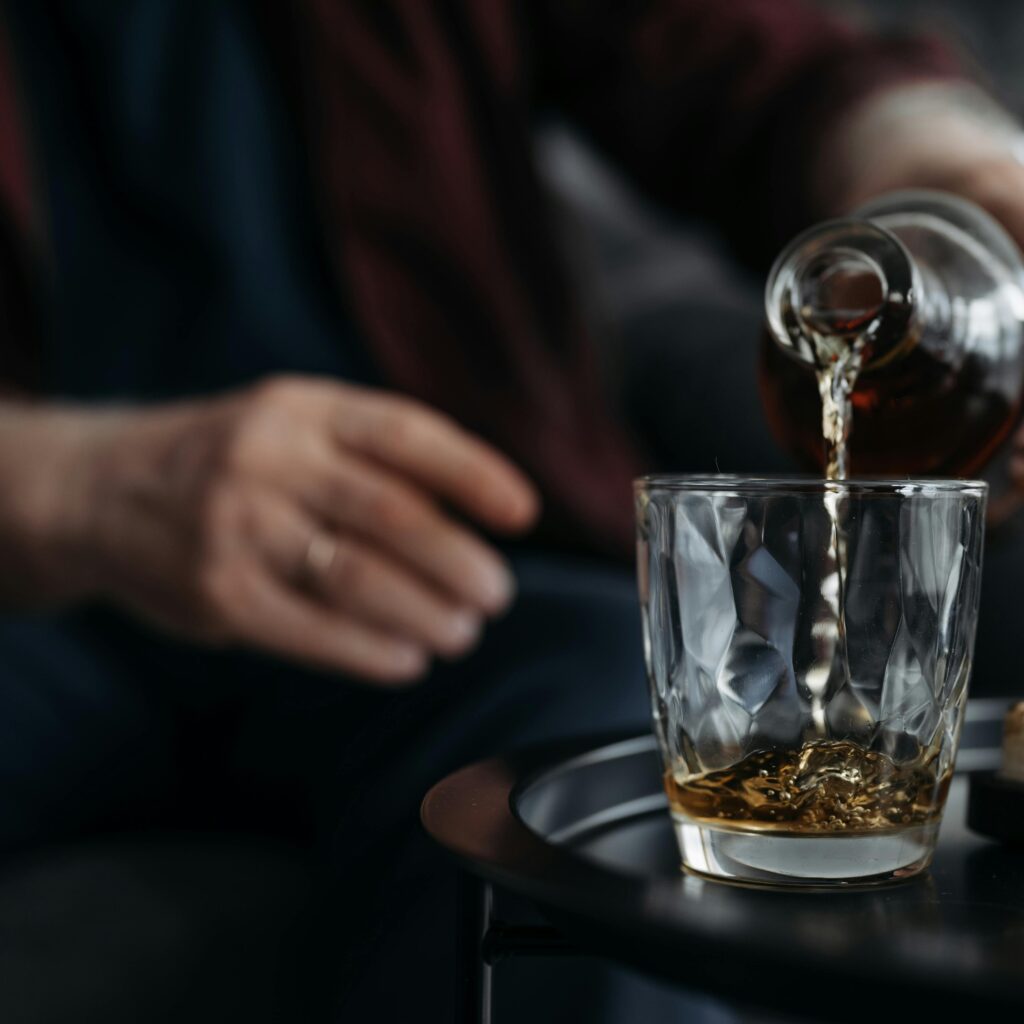 An adult pours whiskey from a bottle into a glass on a table. Indoors setting.