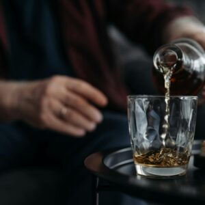 An adult pours whiskey from a bottle into a glass on a table. Indoors setting.